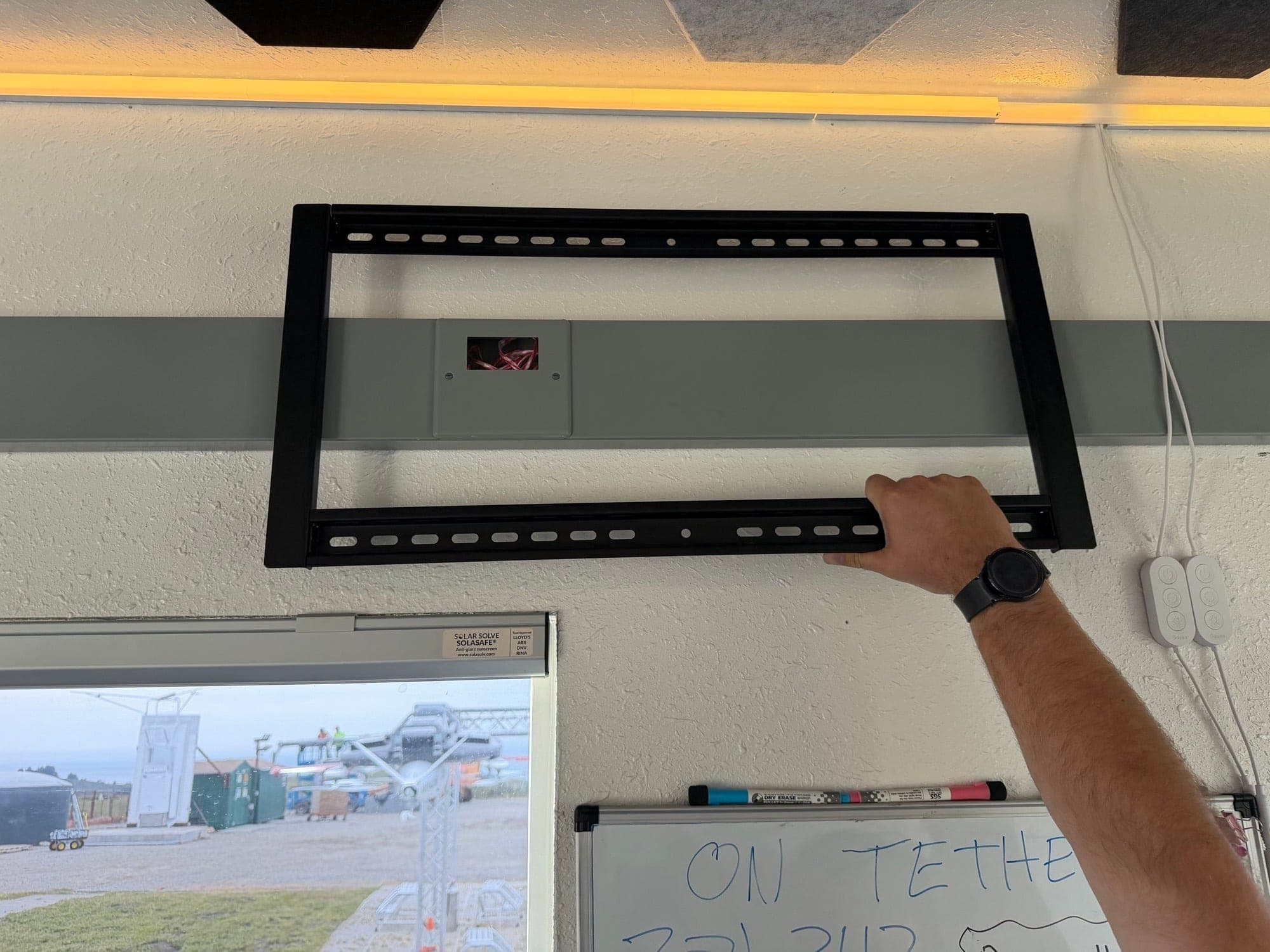 An installer holding a wide TV bracket flat against an office wall next to a recessed outlet plate