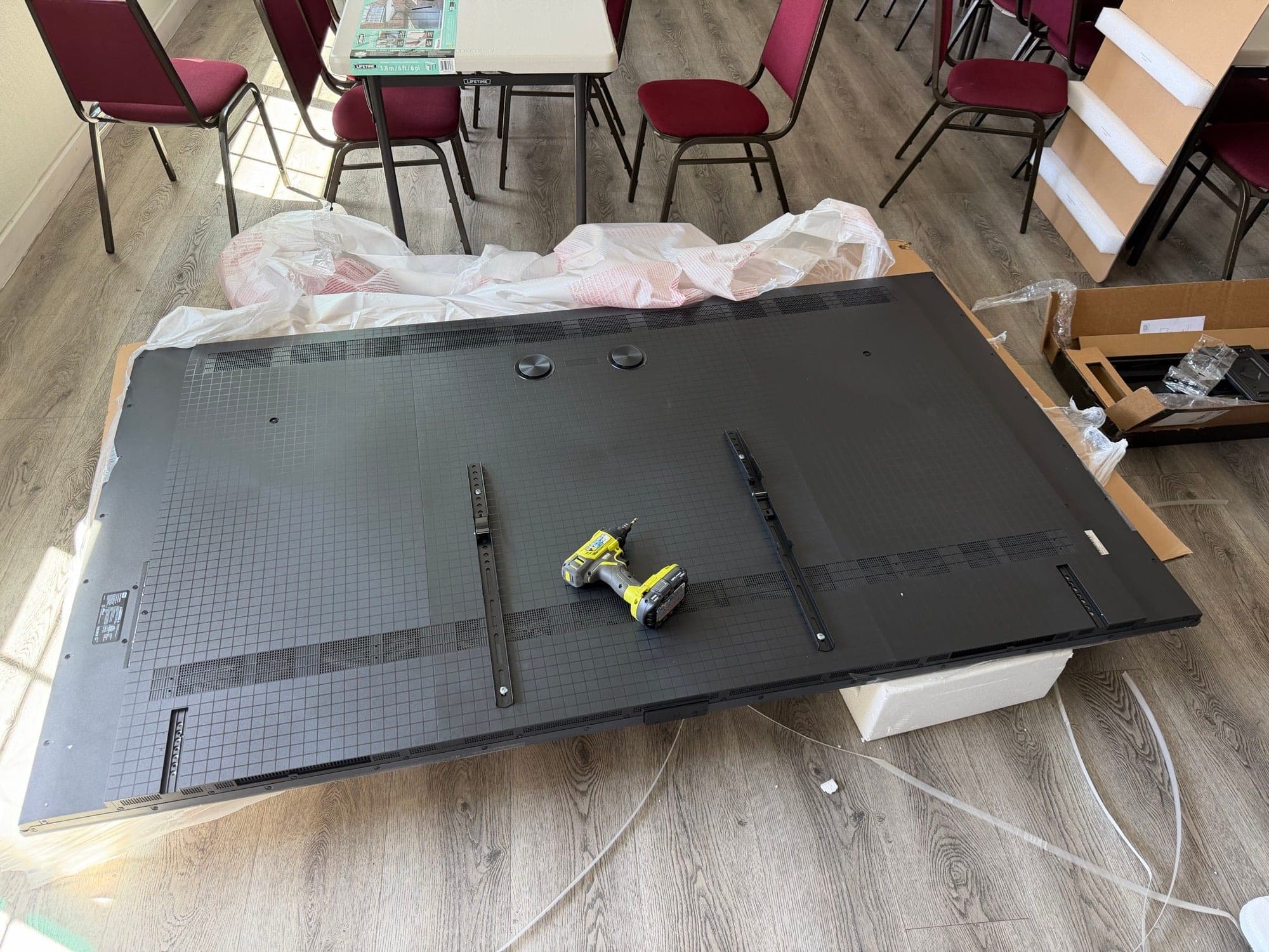 A very large flat panel display face down on packing material with mounting rails being fitted to the back