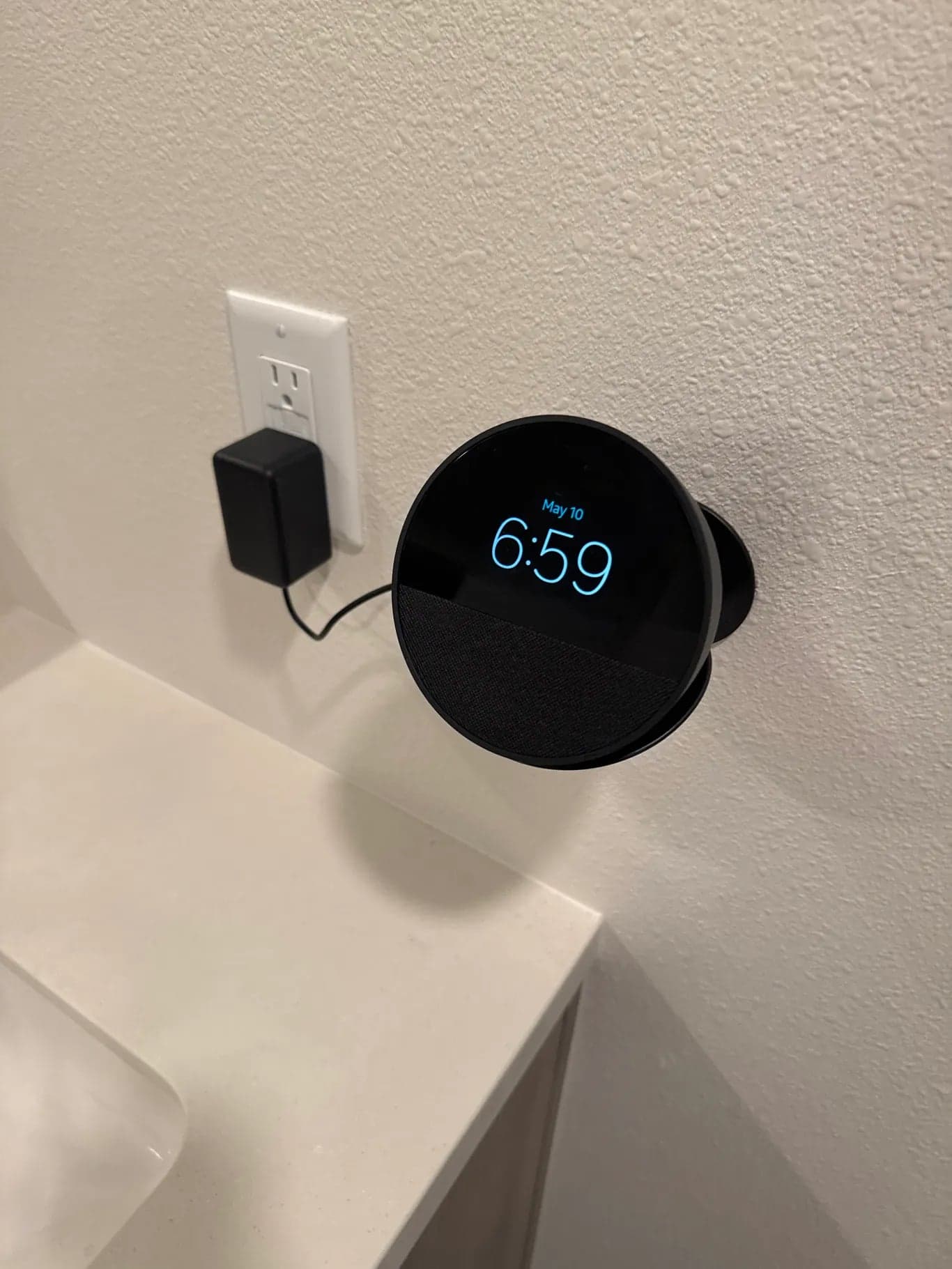 A smart speaker with a clock display mounted on the wall above a bathroom counter