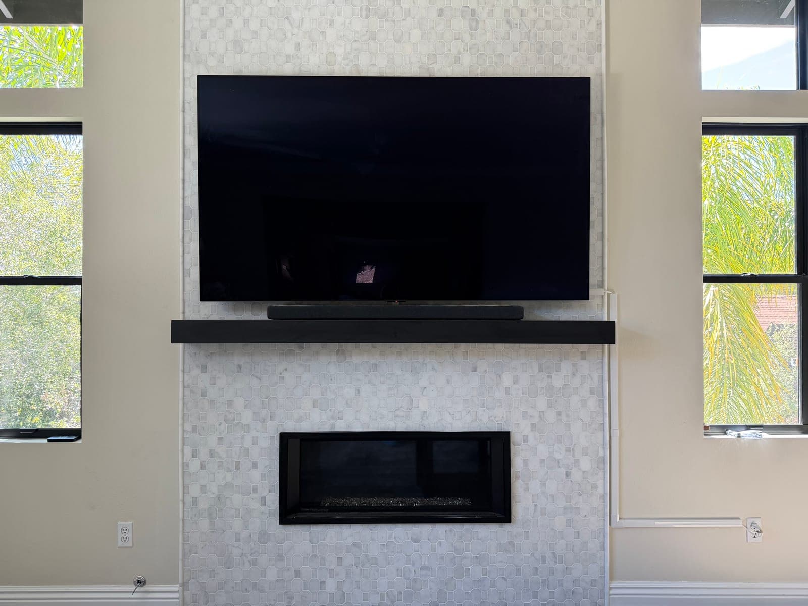 A flat screen TV mounted above a white marble mosaic tile fireplace with a floating shelf and soundbar