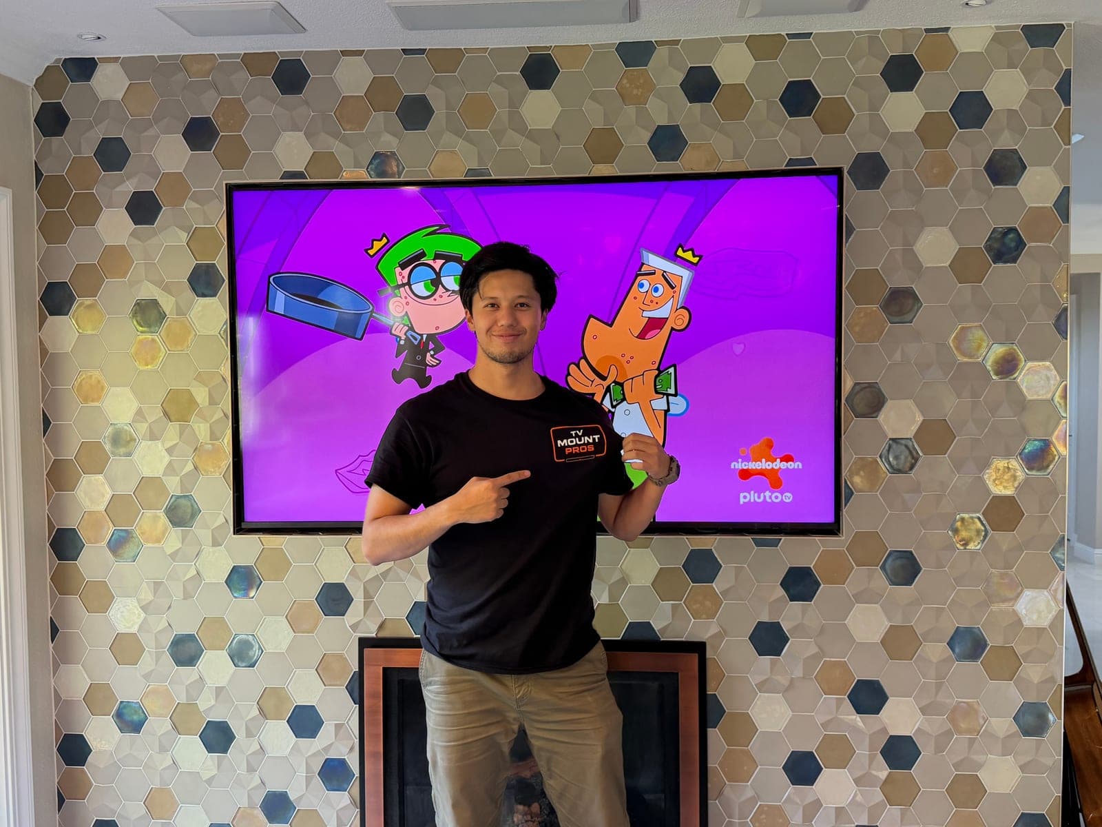 A team member pointing at the TV MountPros logo on his shirt, in front of a TV mounted on a hexagonal tile fireplace wall